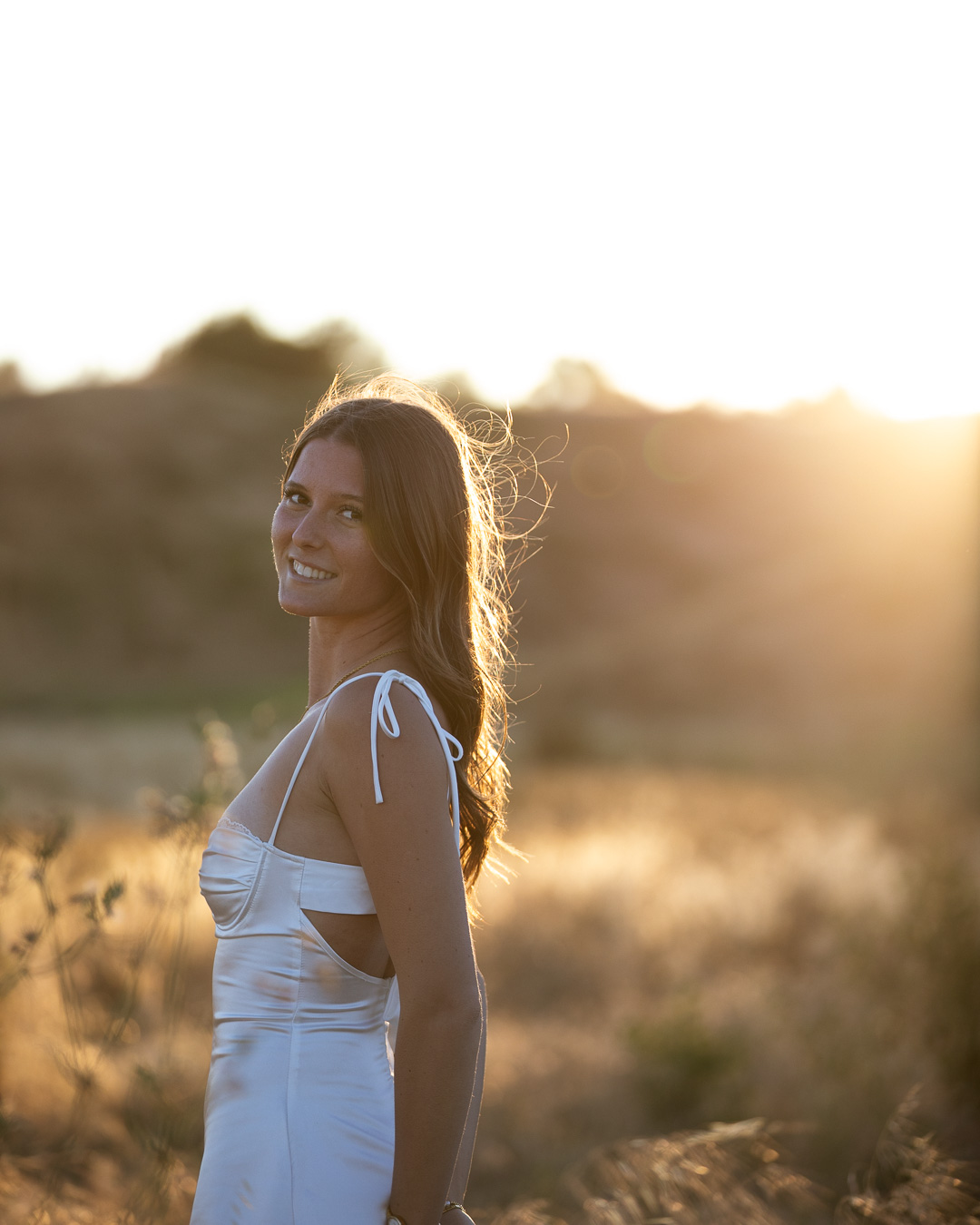 Backlit portrait of a woman in a white satin dress smiling over her shoulder at sunset